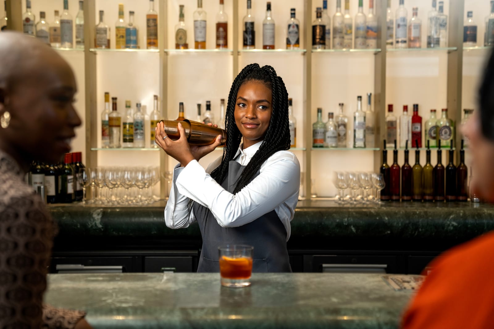 A bartender shaking a cocktail behind a warmly lit bar while guests chat